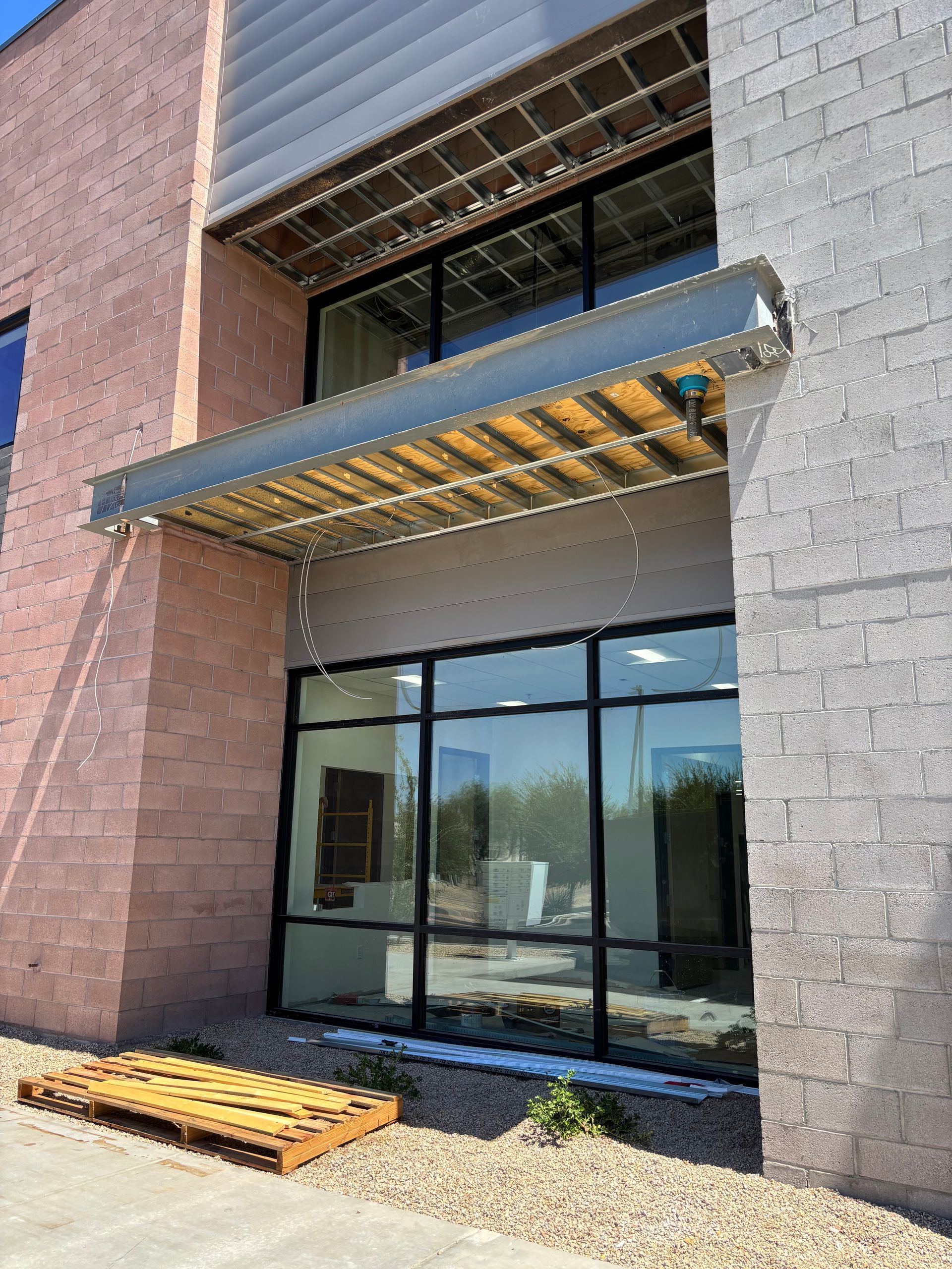Exterior view of a commercial building with large windows and a metal awning. A wooden pallet sits on the ground.