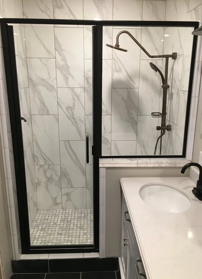 Bathroom with marble-patterned tile shower, black-framed glass door, bronze shower fixtures, and white vanity.