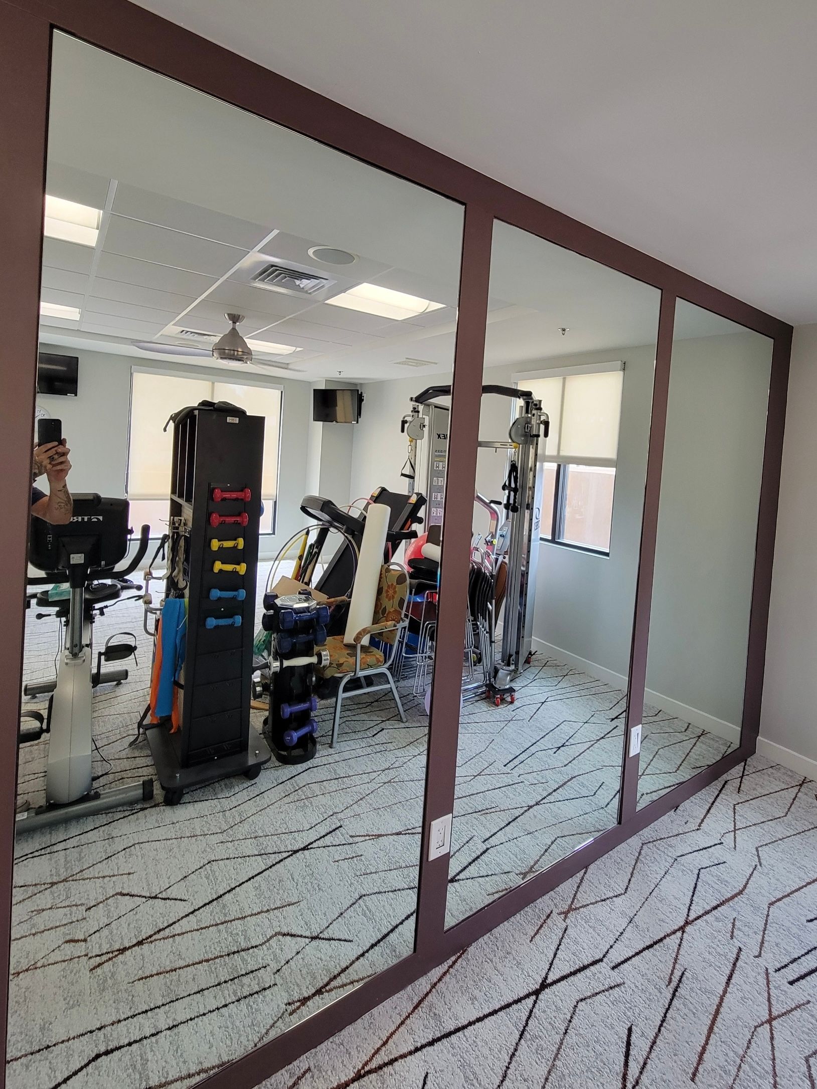Gym interior with large mirrors reflecting exercise equipment and a ceiling fan.