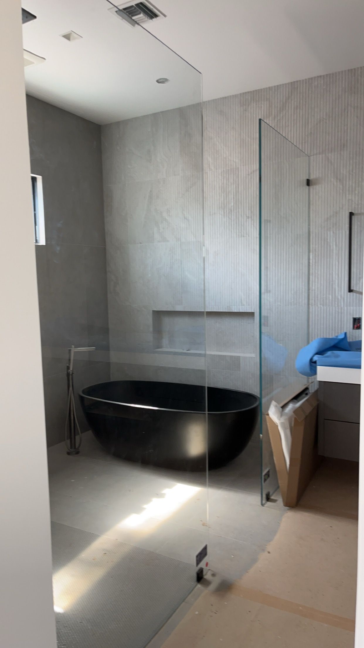 Modern bathroom with gray tile walls, black tub, glass shower enclosure, and sunlight.