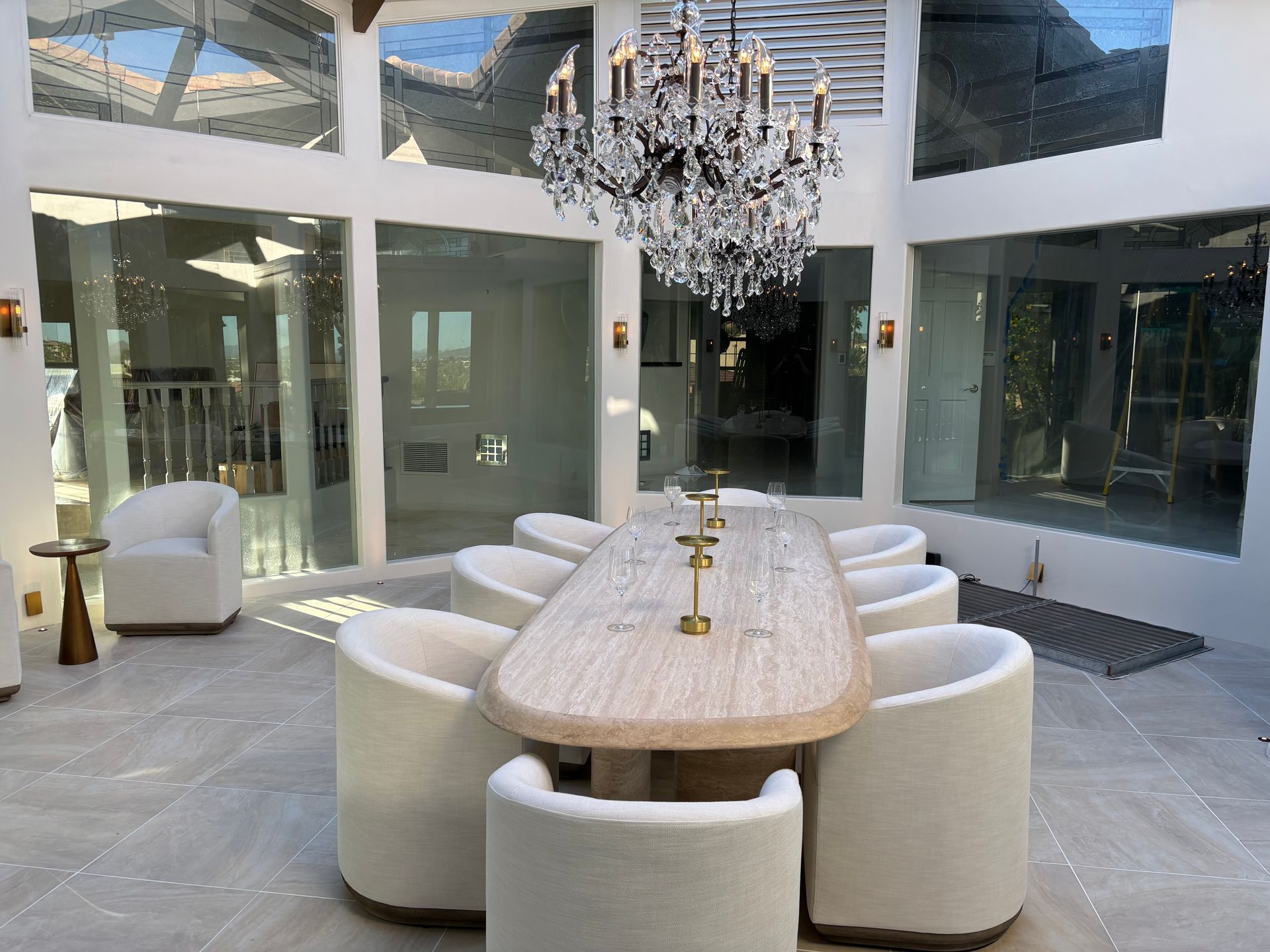 Dining room with a long table, white chairs, and a chandelier, surrounded by windows.