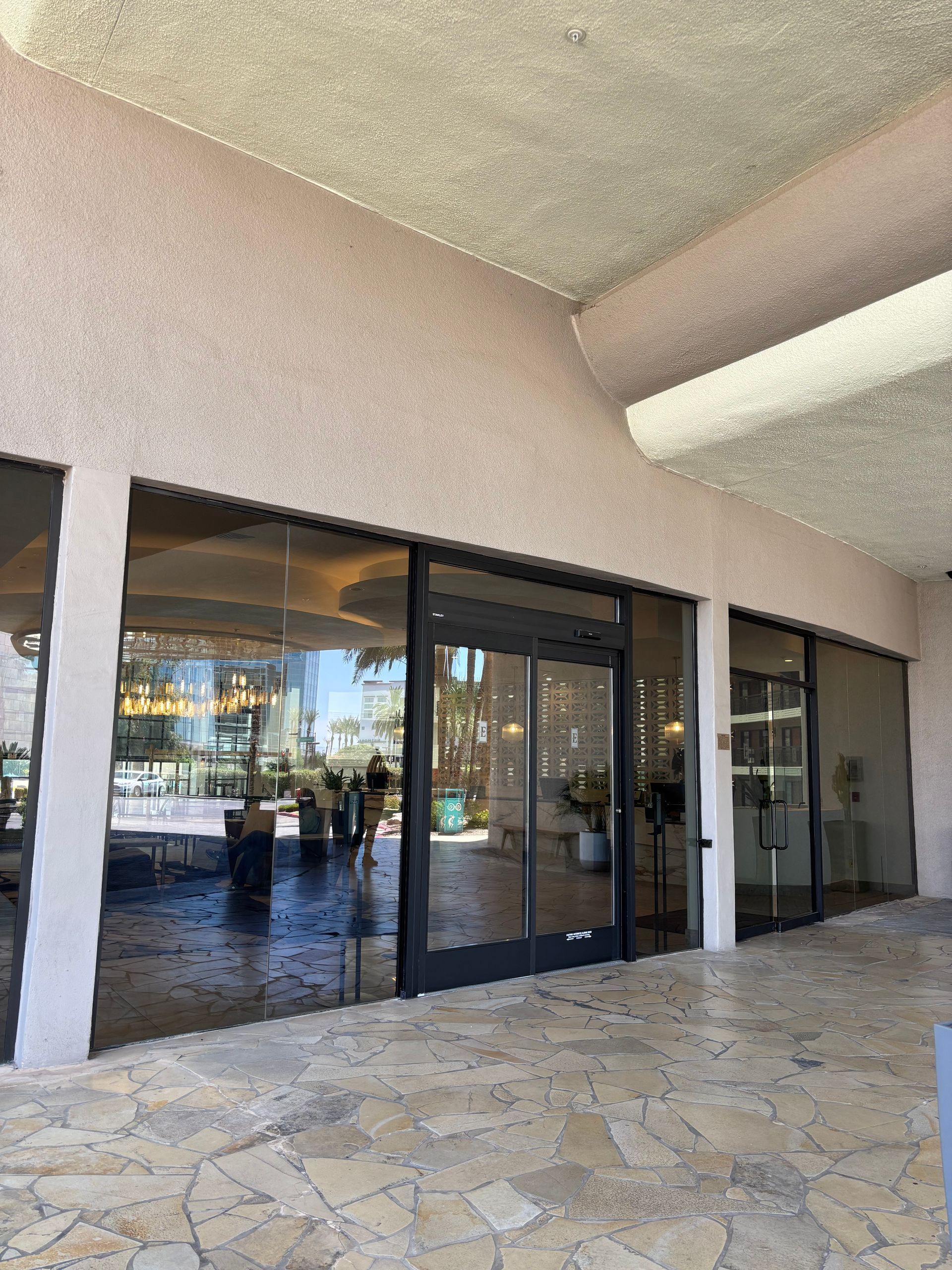 Exterior of a building with large glass windows and doors, under a covered walkway, paved ground.