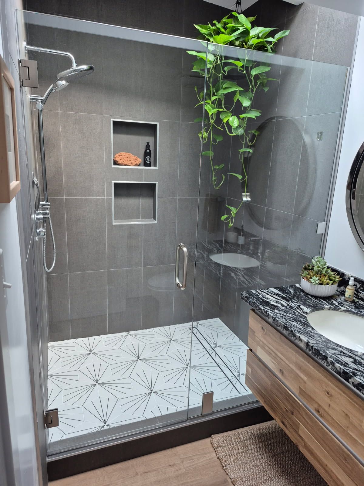 Modern walk-in shower with gray tile, glass door, white patterned floor, and a built-in niche with plants.