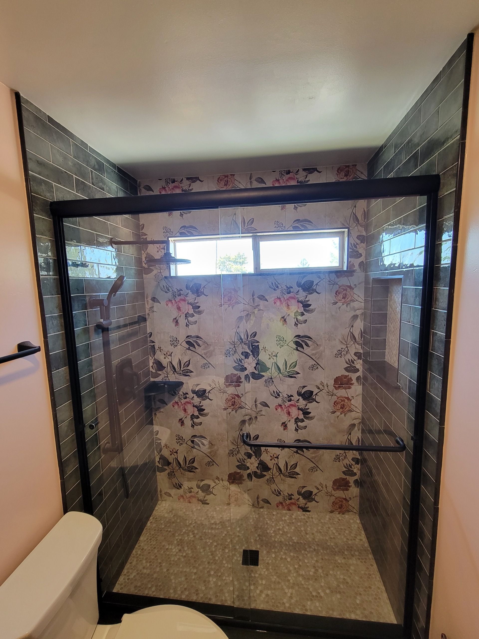Shower with floral wallpaper, black-framed glass door, gray tile, and black fixtures.