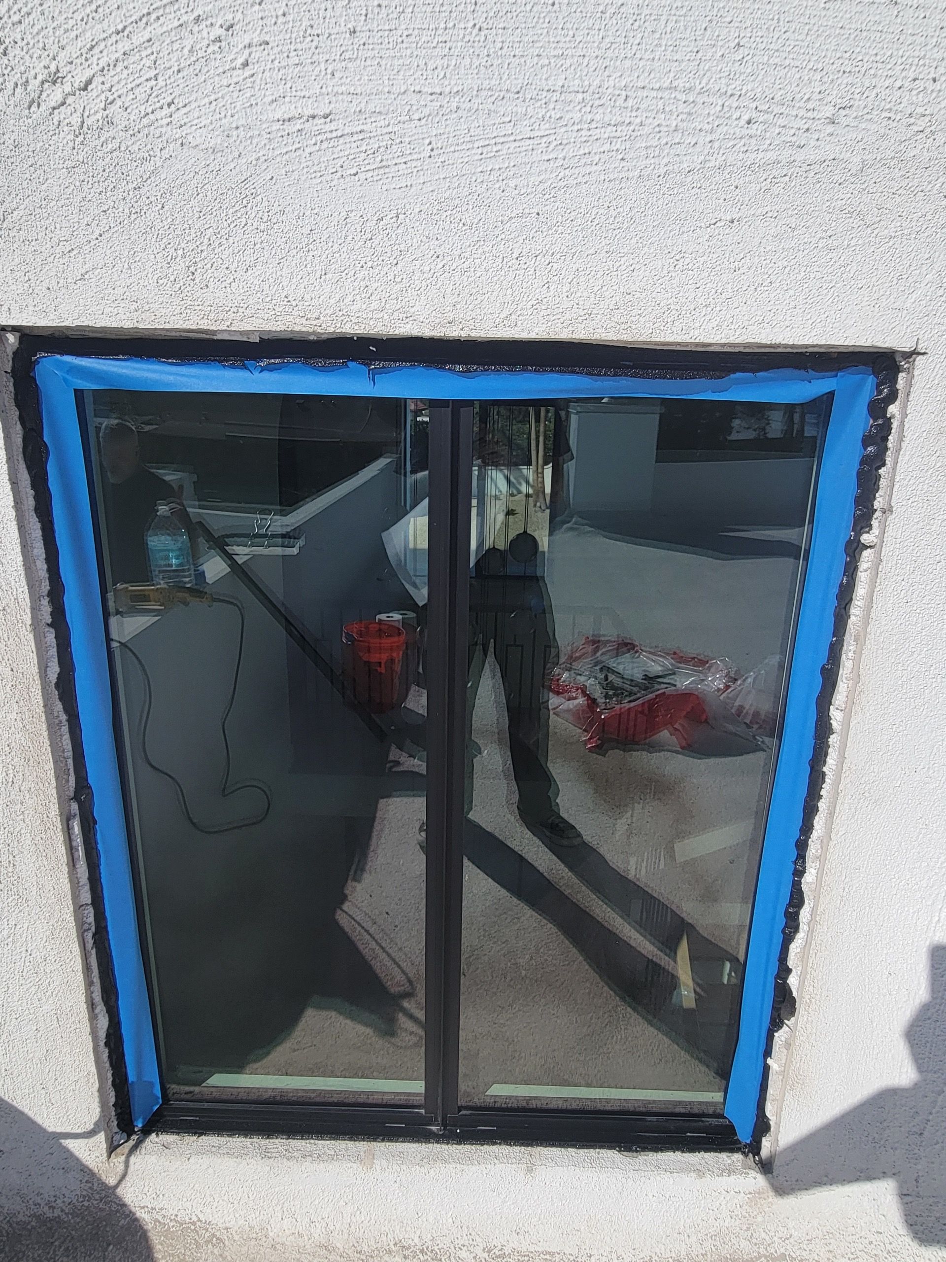 Window with blue tape around the edges, reflecting a person and outdoor setting.