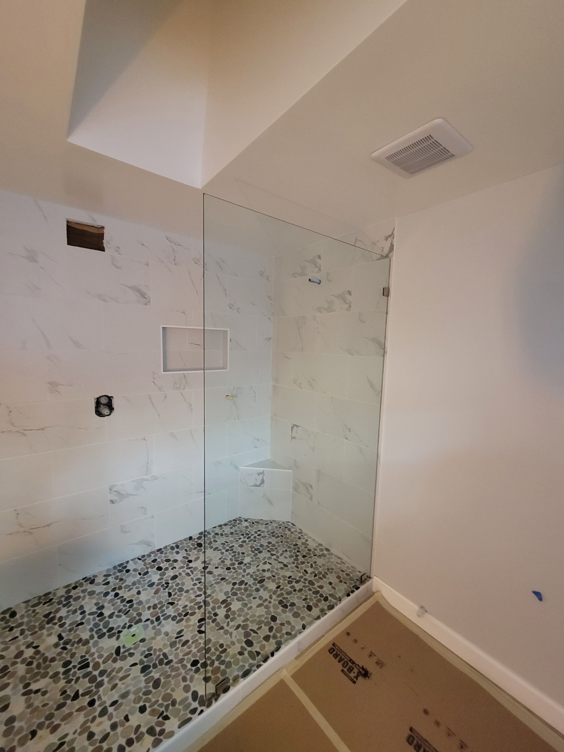 Shower with clear glass walls, pebble floor, and white marble-patterned tile, with a white ceiling and vent.