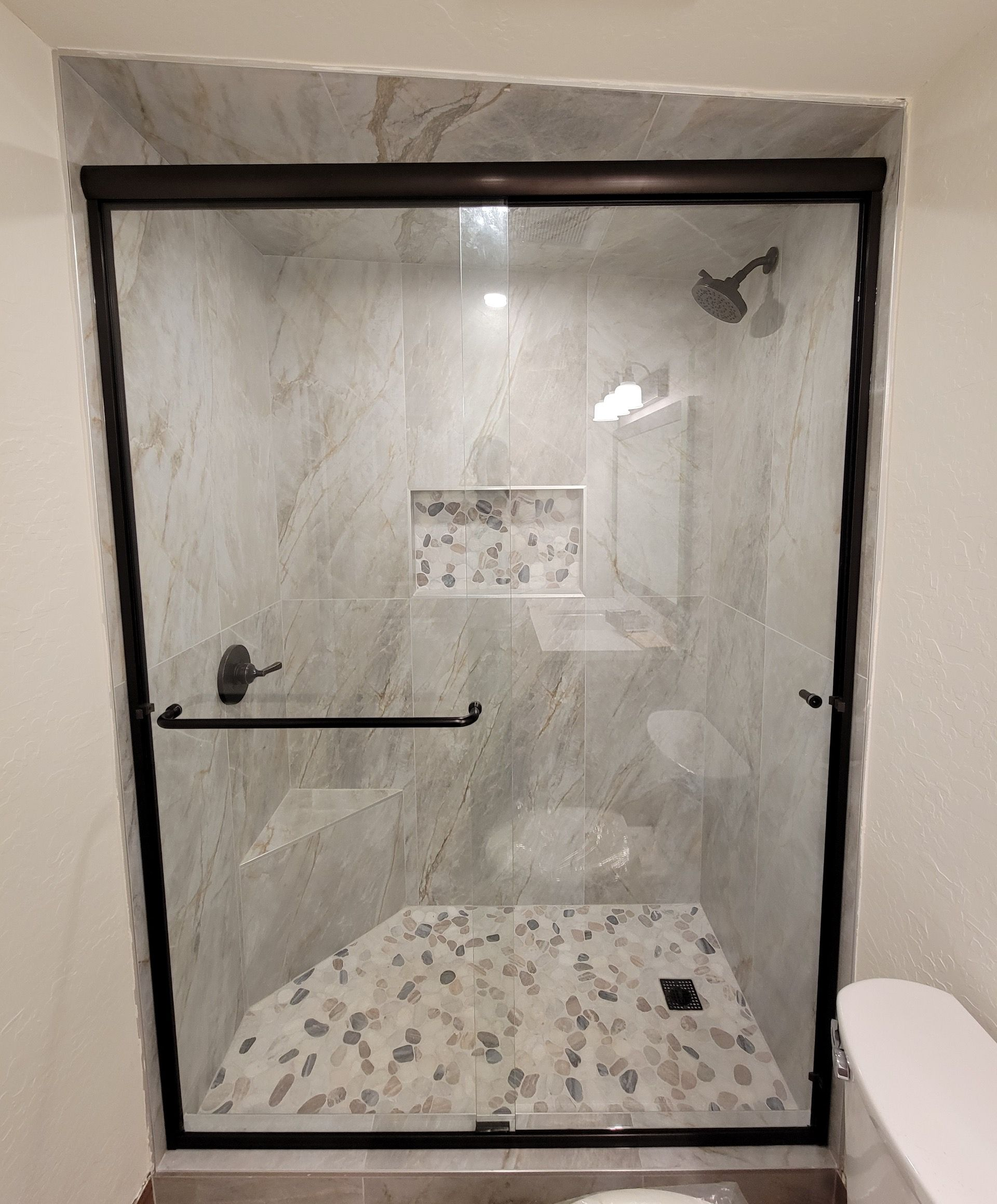 Black framed glass shower with stone floor and tile walls. A built-in shower niche with a light above.