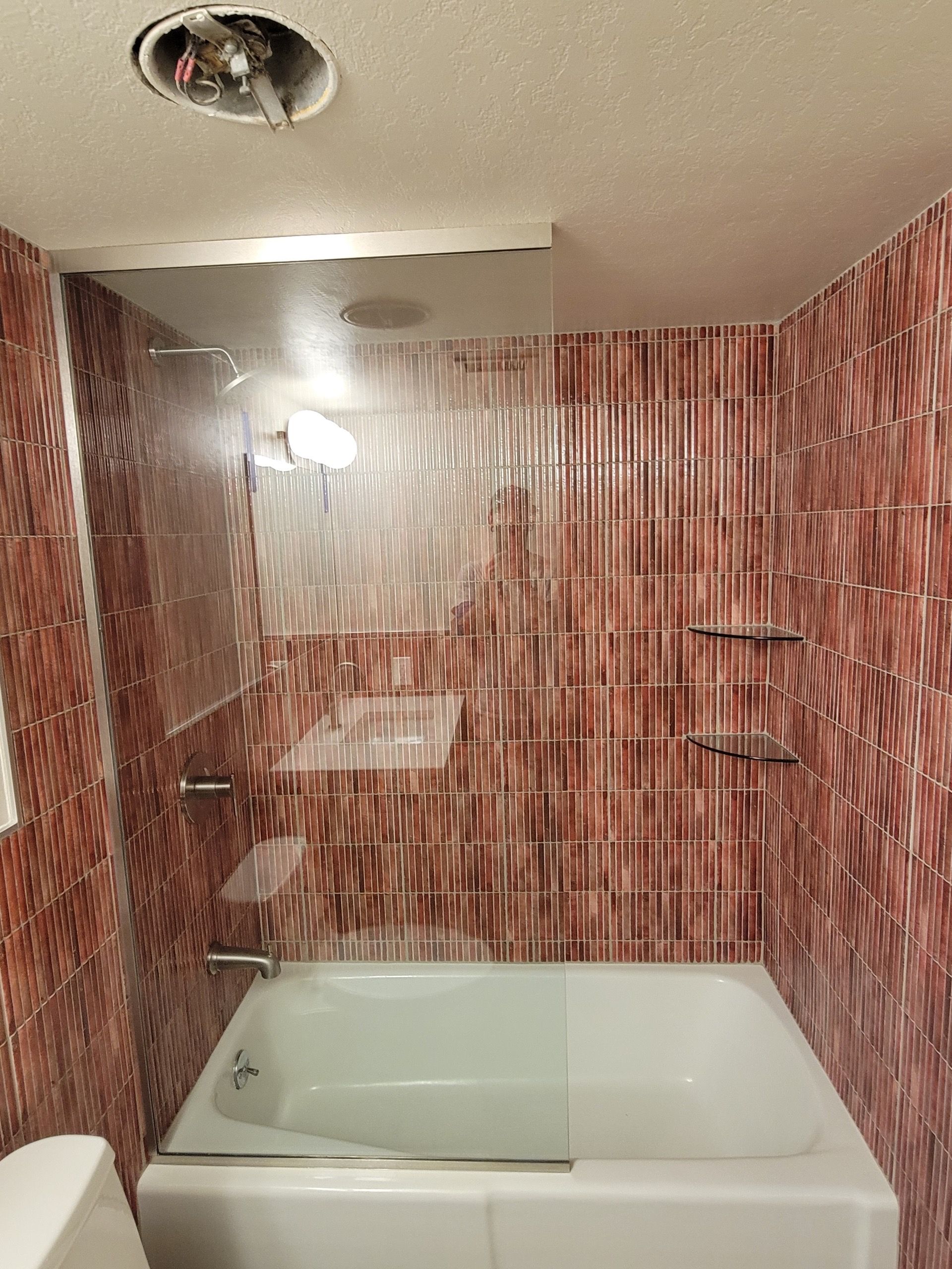 Bathroom with red-brown tile walls, white bathtub, glass shower screen, and a visible ceiling light fixture.