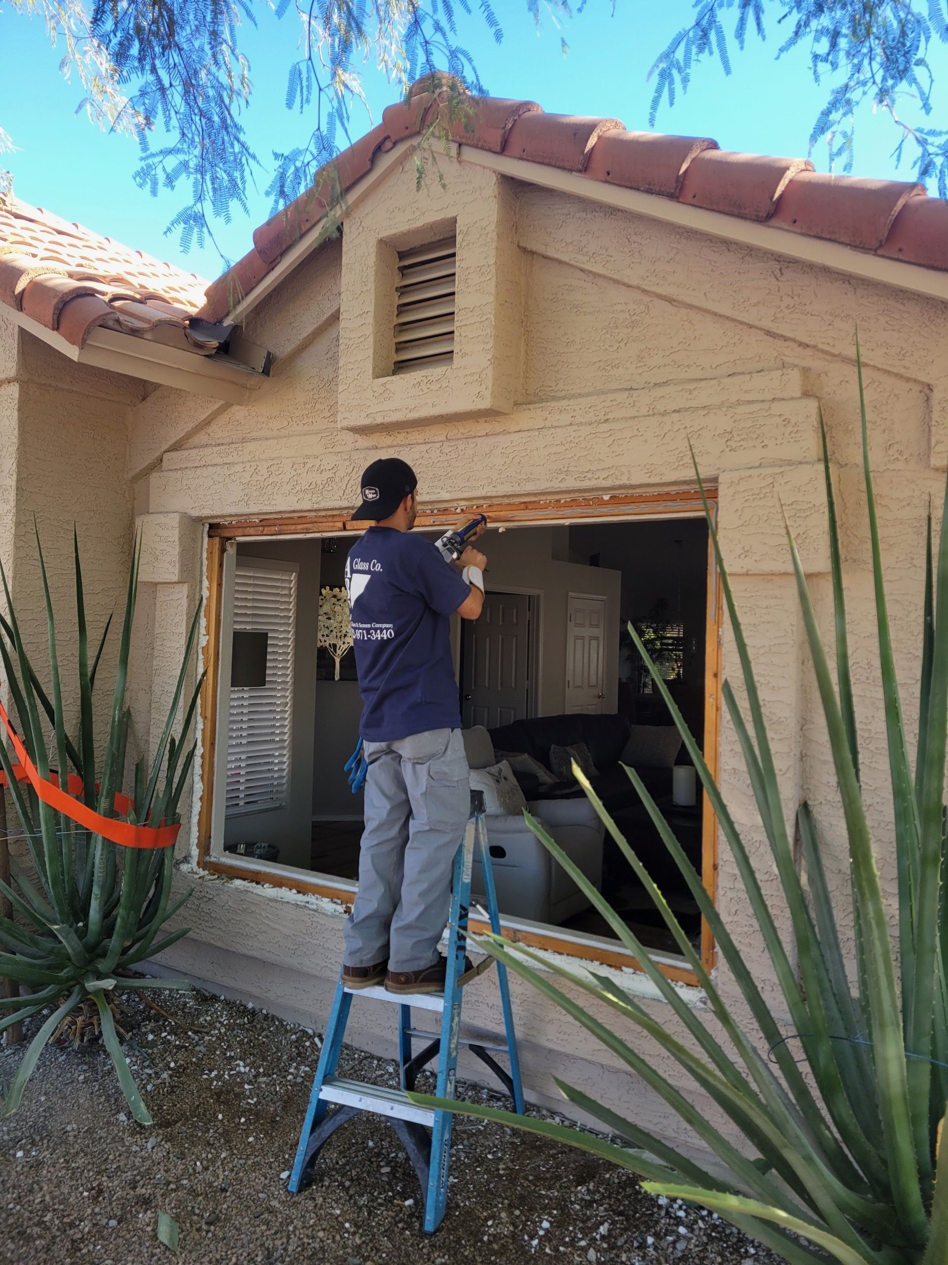 A person on a ladder repairs a window on a house. The house has a tan exterior and red tile roof.