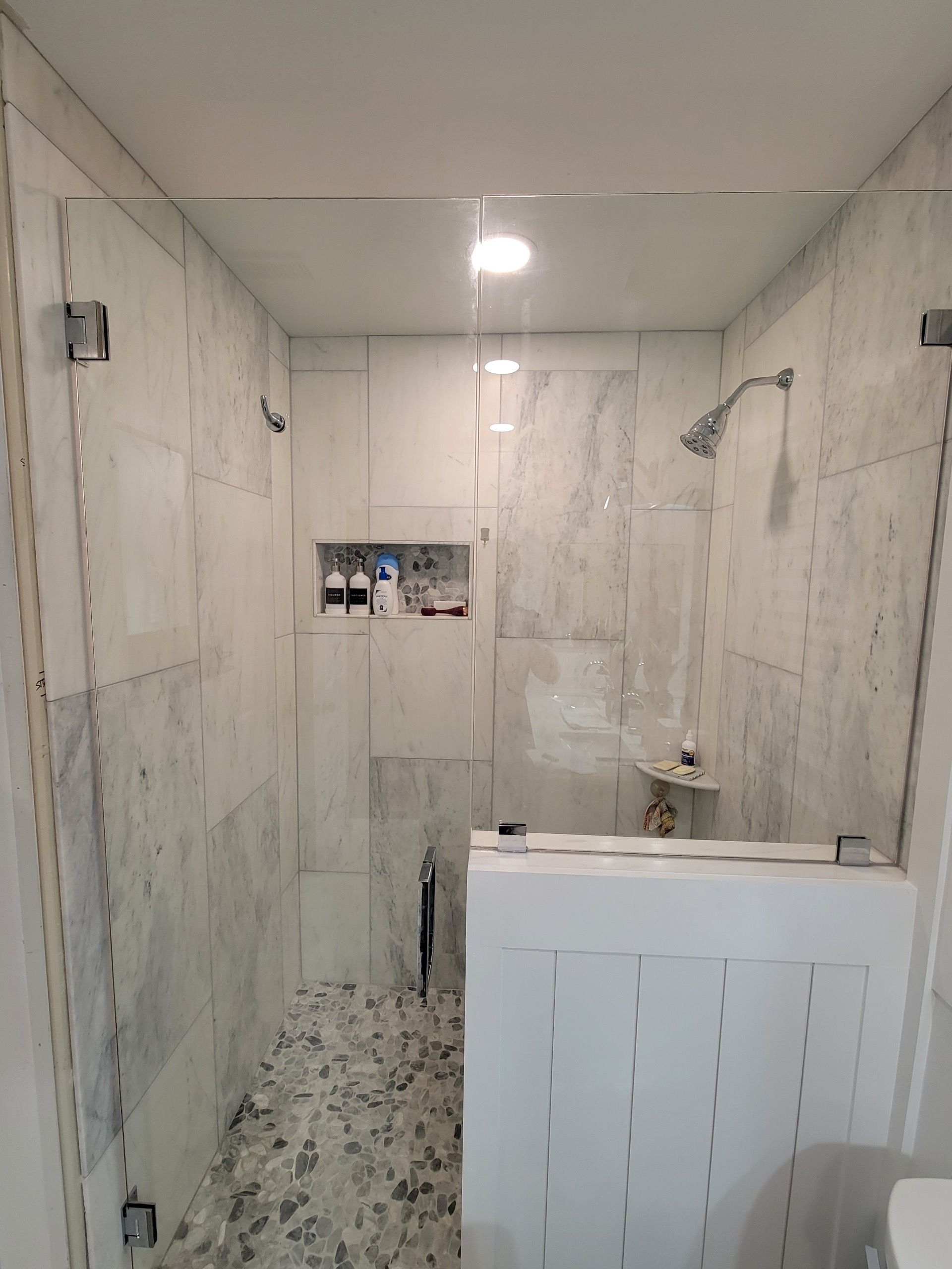 Bathroom shower with glass doors, marble tiles, pebble floor, and white accent wall.