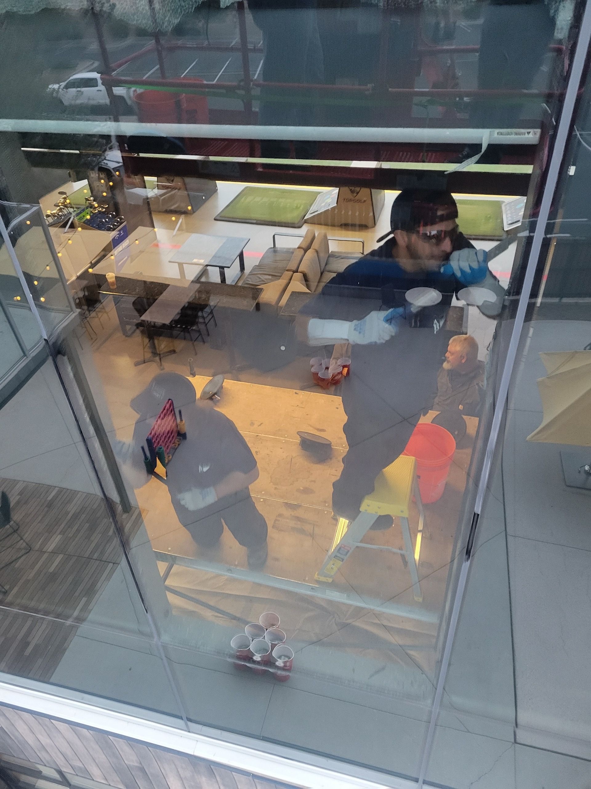 Two people cleaning a large glass window of a building; reflection shows outdoor patio furniture.