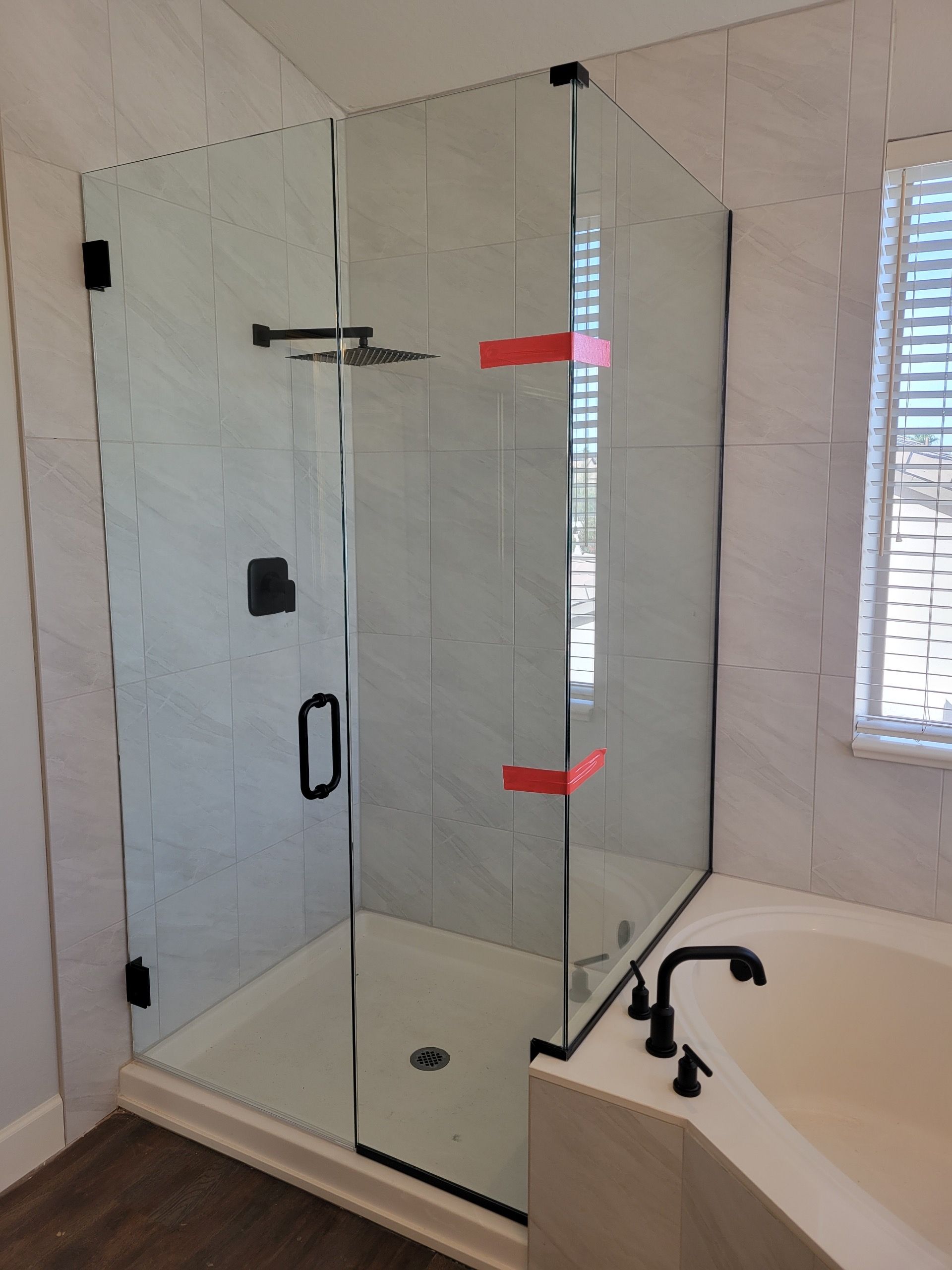 Glass shower enclosure with black fixtures and red support pieces next to a white bathtub.