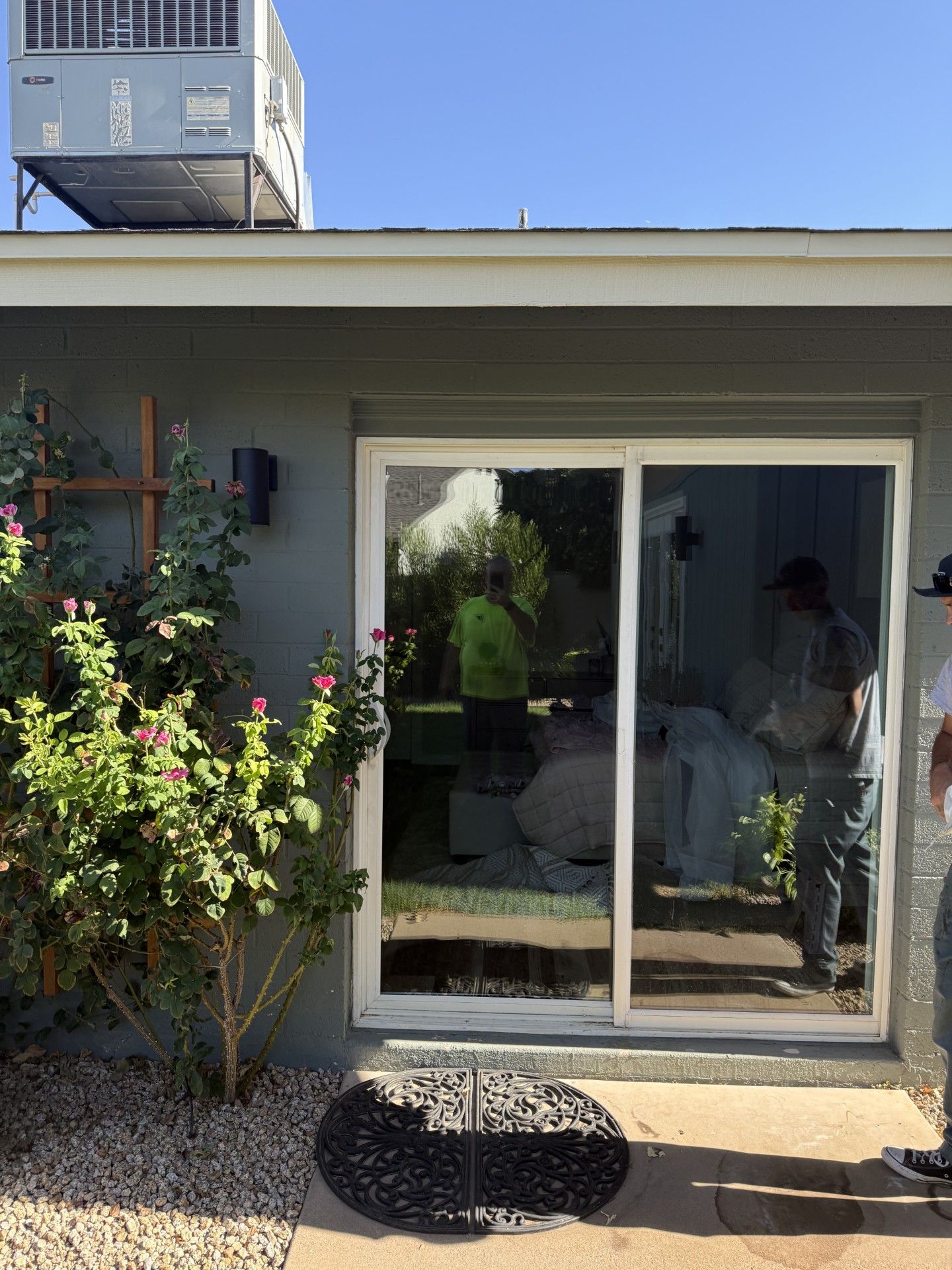Sliding glass door reflects workers, green shirt. Building has gray walls, bushes, and pebble ground.