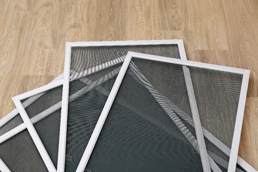 White-framed window screens stacked on a light wooden surface, with dark gray mesh.