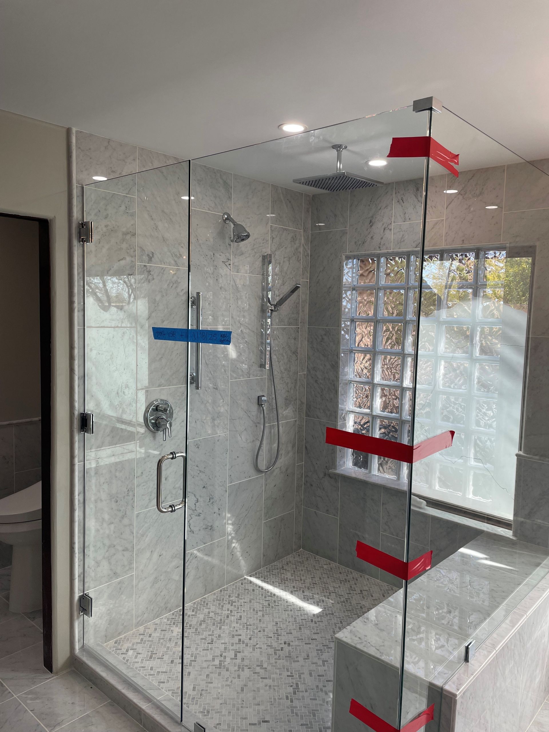 Glass shower enclosure with gray tiled walls and floor. Red and blue protective tape on glass.