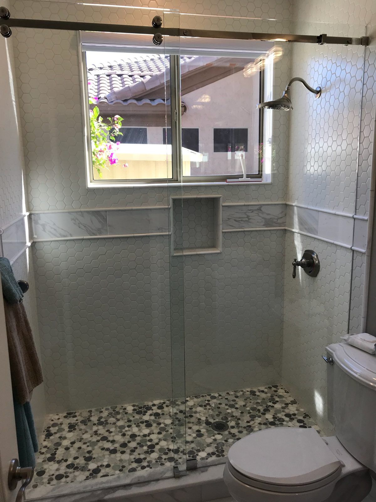 Sliding shower glass with dark bronze roller hardware under a window with pebble shower floor in Phoenix AZ