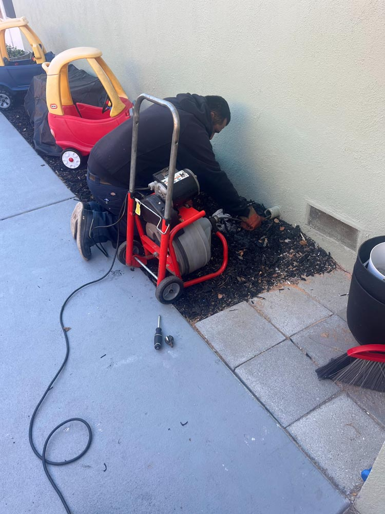 Worker using a red floor machine to clean debris on a sidewalk beside a wall