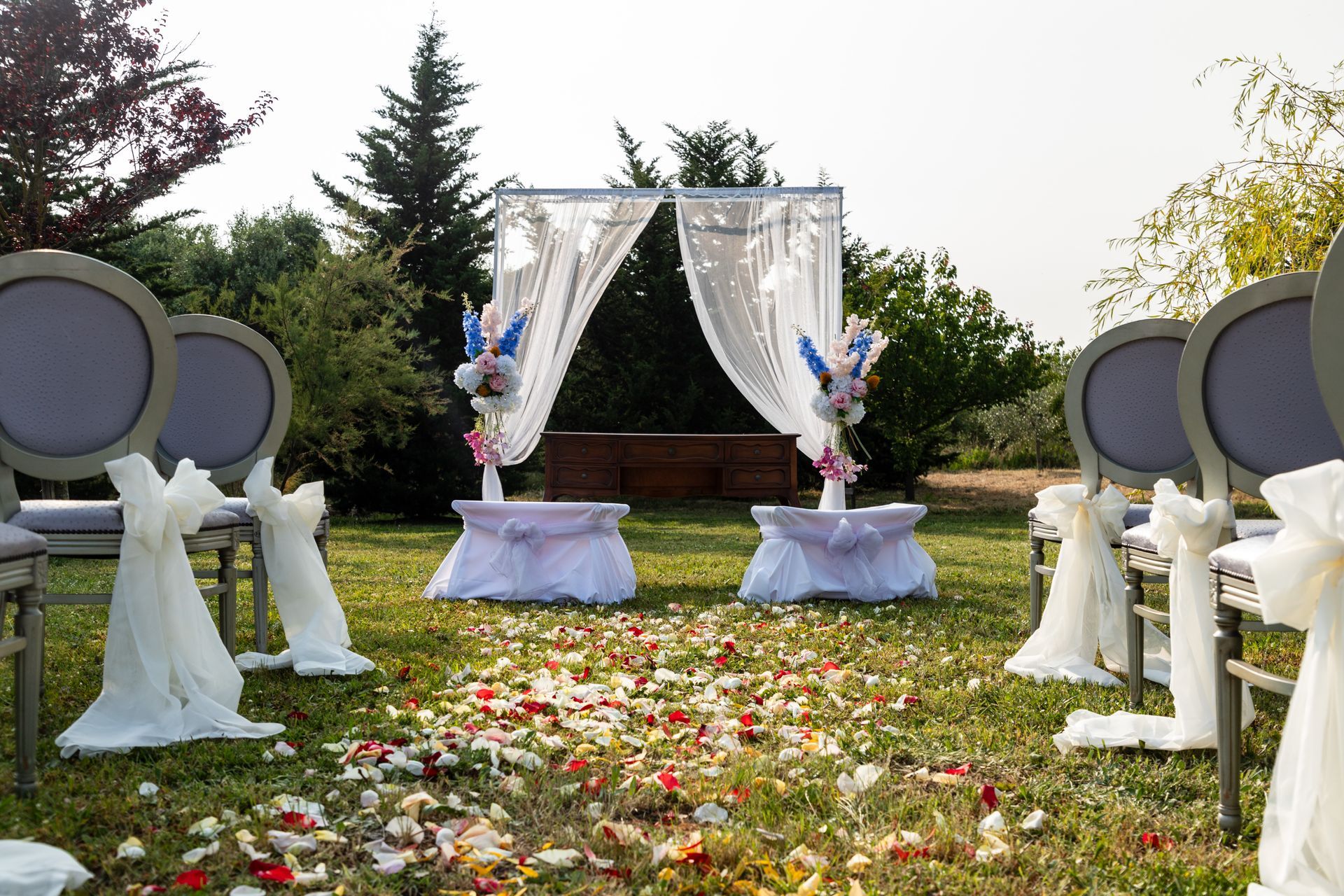 Navata nuziale con arco drappeggiato di bianco, petali di fiori e sedie rotonde all'aperto in un giardino.