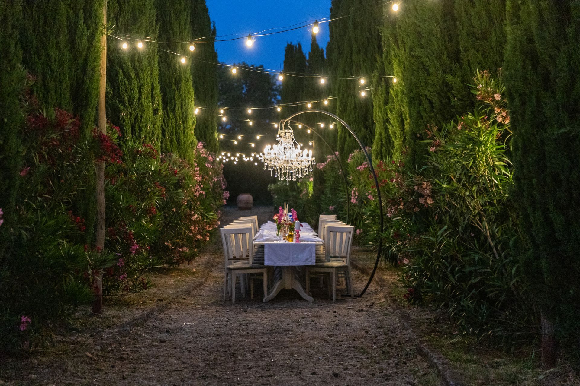 Un tavolo da pranzo all'aperto in un lussureggiante sentiero del giardino, illuminato al crepuscolo da lucine e lanterne.