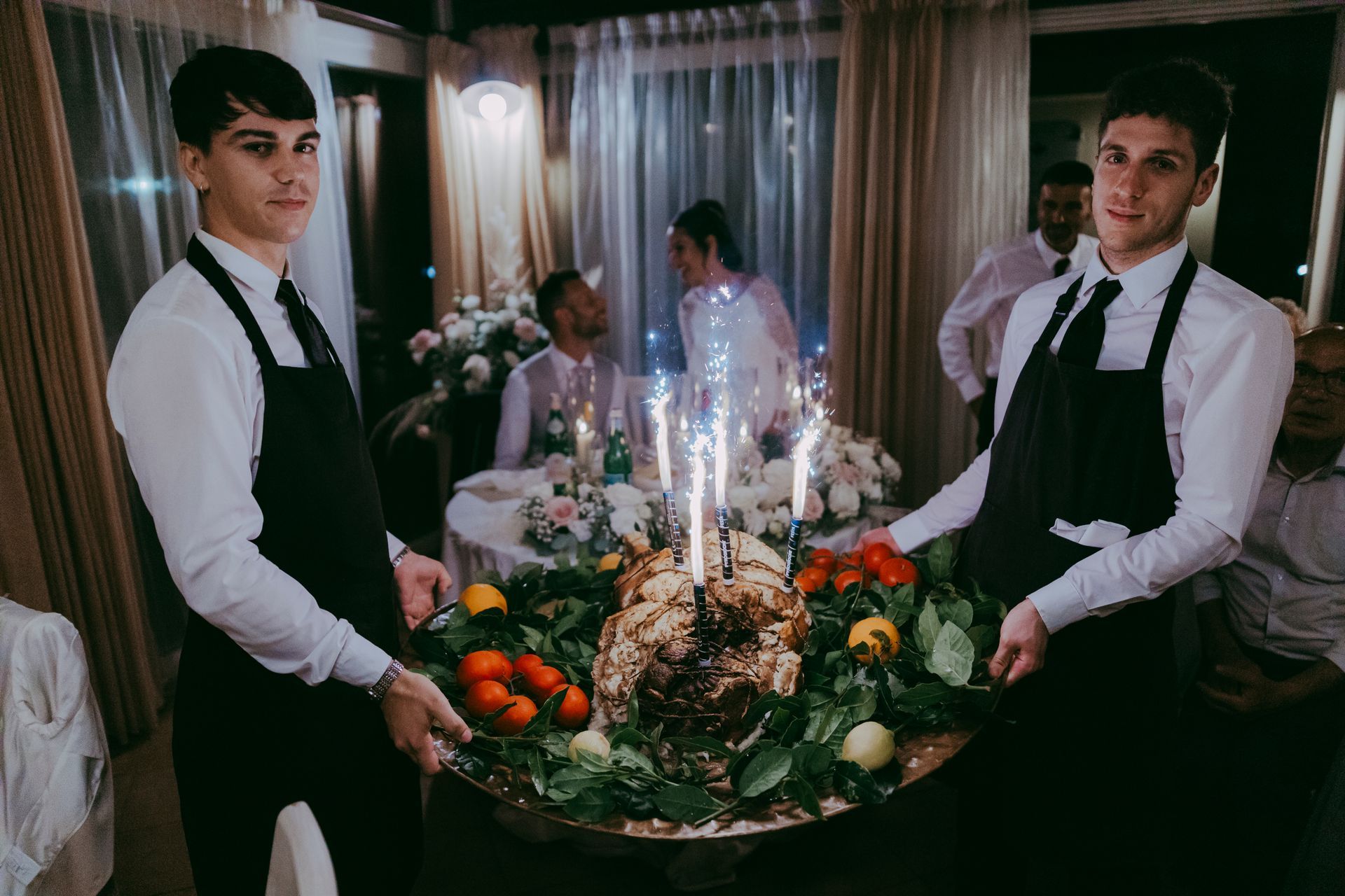 Due camerieri che portano un grande centrotavola floreale con candele accese a un tavolo da pranzo formale.