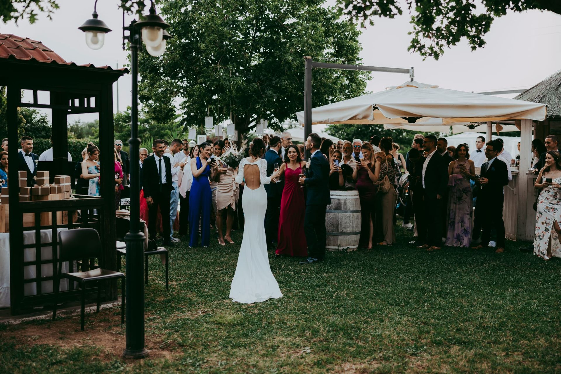 La sposa, in abito bianco, si rivolge alla folla durante un ricevimento di nozze all'aperto, sotto tendoni e alberi.