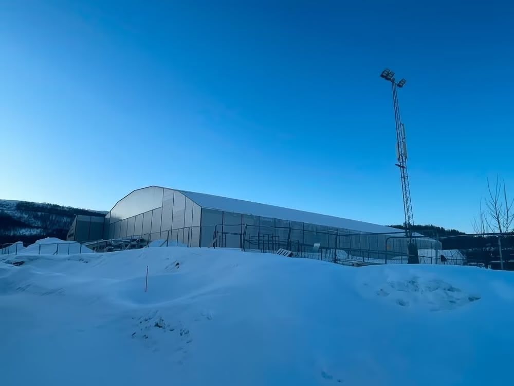 Snow-covered landscape with a large white building and tall light pole under a clear blue sky.