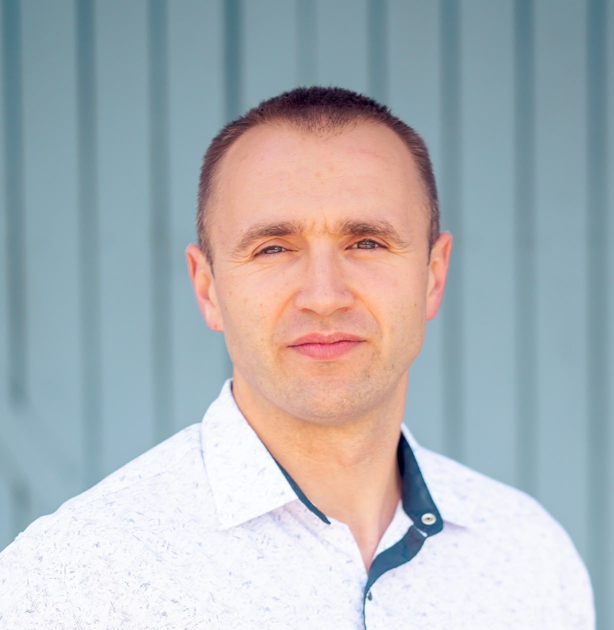 Man in white patterned shirt smiles slightly, standing against a blue wall.