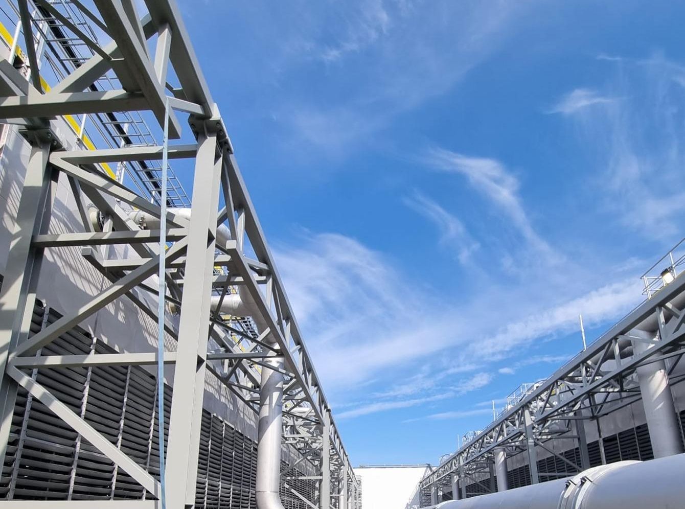 Metal framework of industrial structures against a bright blue sky with wispy clouds.
