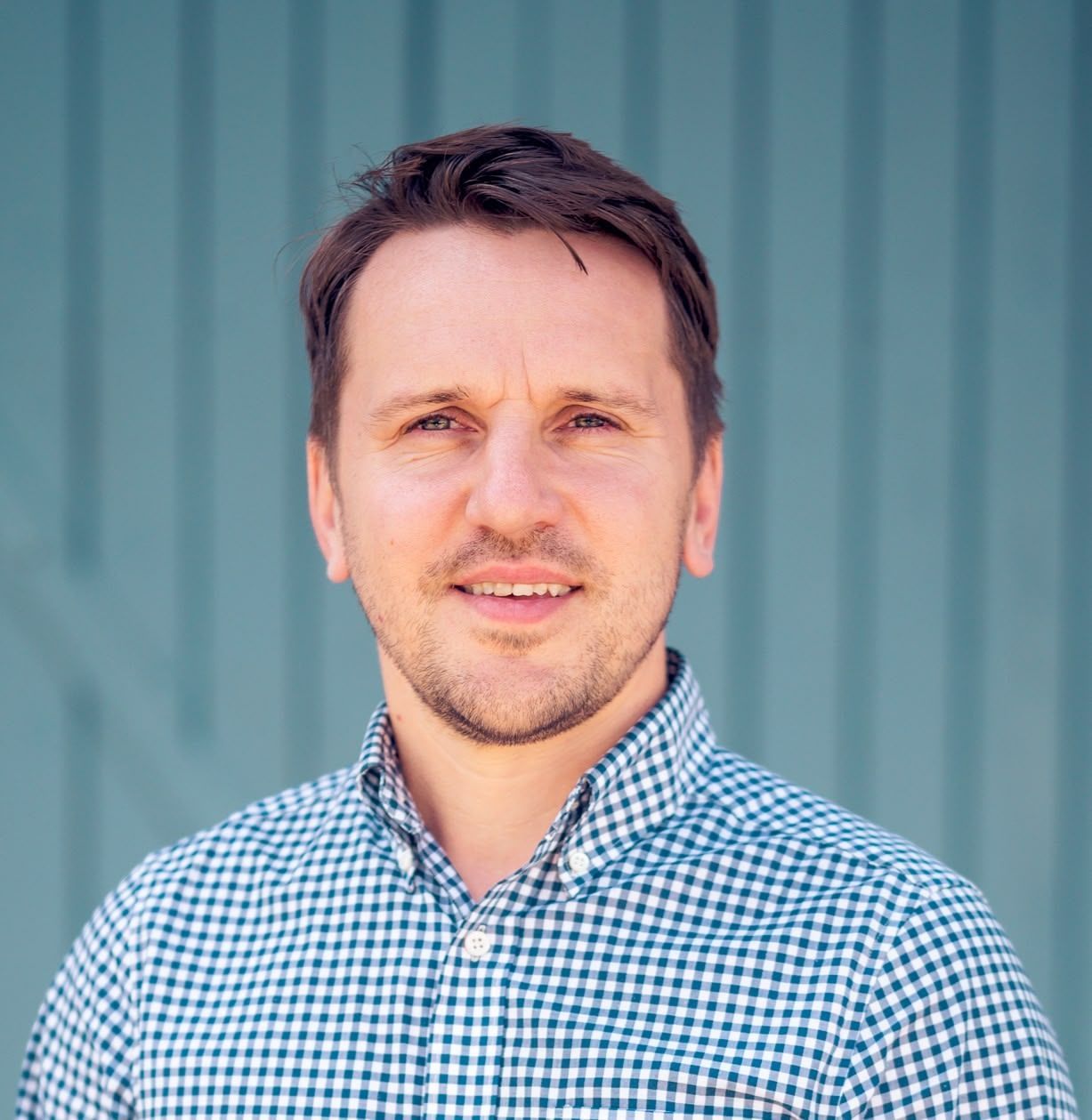 Man in checkered shirt smiles in front of a blue wall.