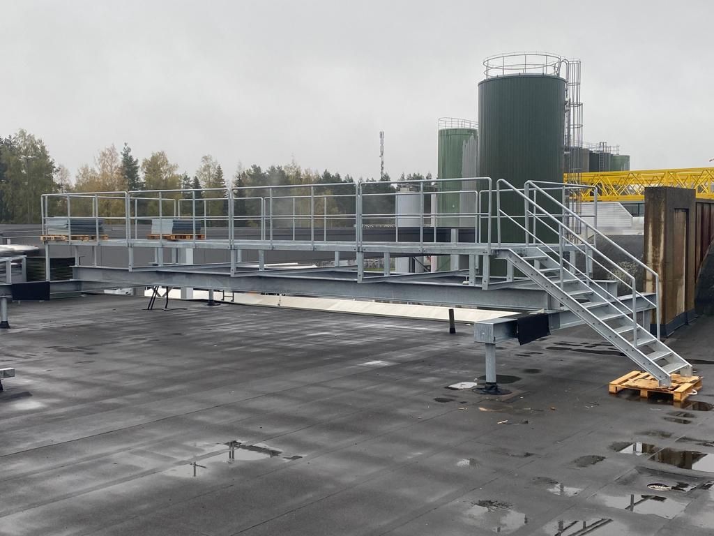 Metal walkway with stairs and railing, industrial setting with storage tanks and overcast sky.