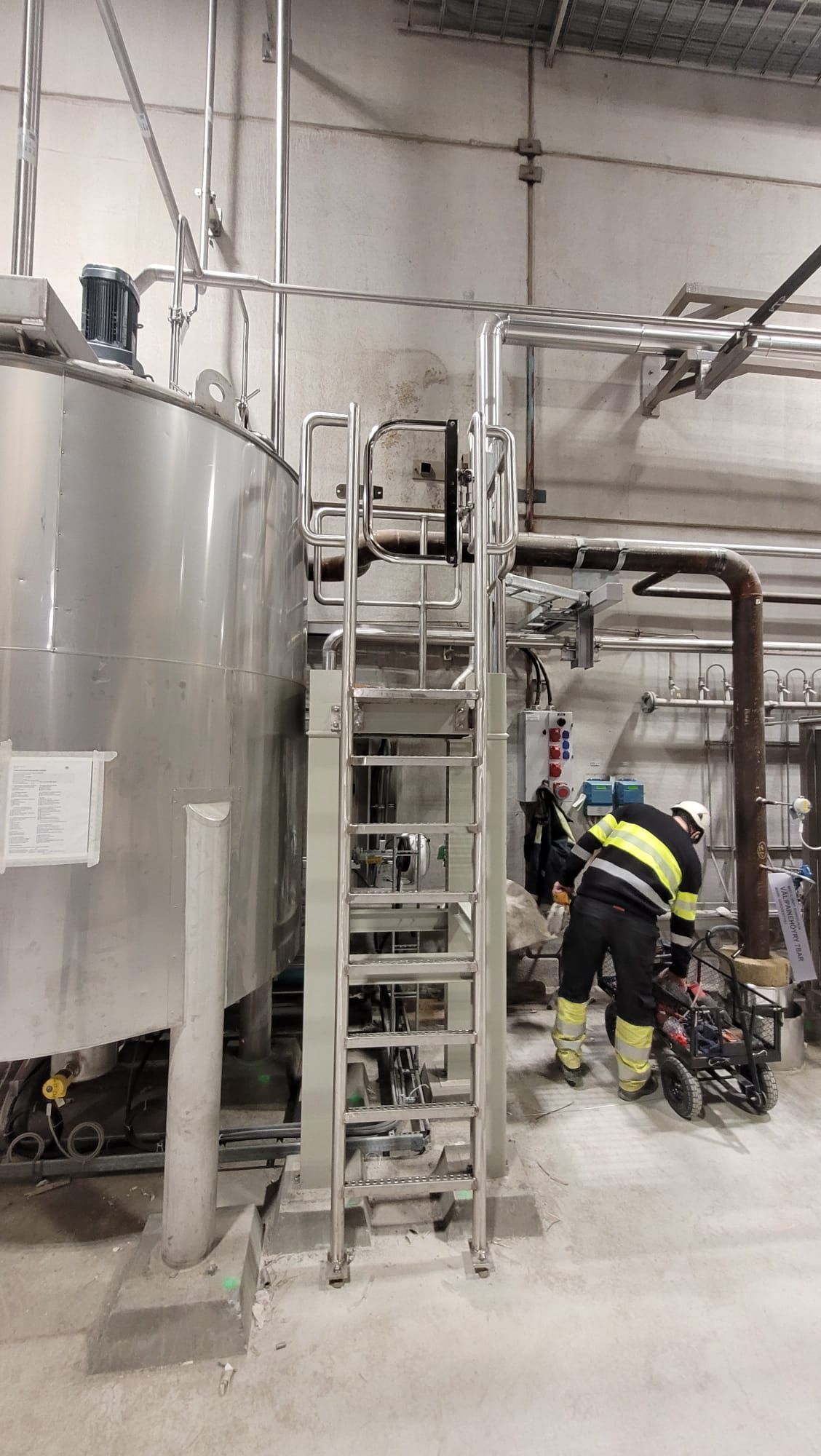 Stainless steel tank with a ladder. A worker in safety gear near pipes. Industrial setting.