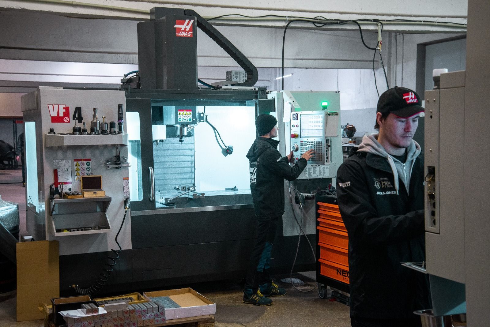 Two workers operate CNC machines in a workshop; one adjusts controls, the other monitors a display.