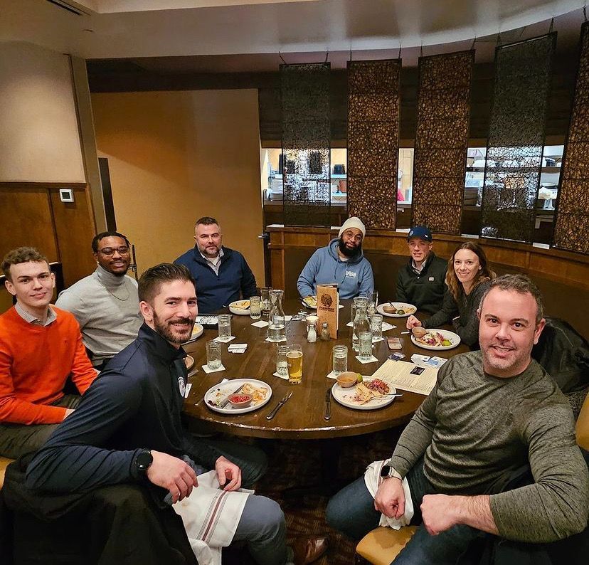 Seven people seated around a restaurant table, smiling and chatting over drinks and plates.