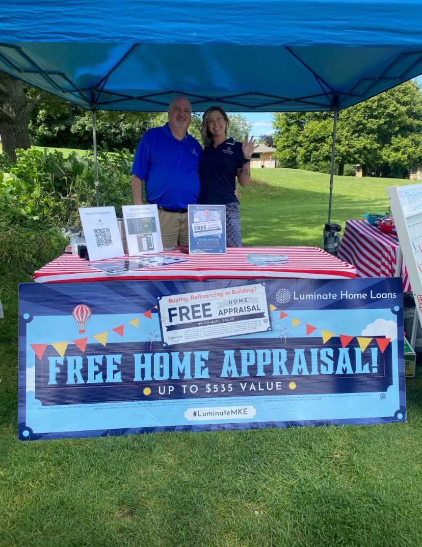 Two people behind a table at a “FREE HOME APPRAISAL!” booth under a blue tent in a grassy park.