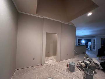 Unfinished room with gray walls, open doorway, ceiling lights, and painting supplies on a dusty floor.