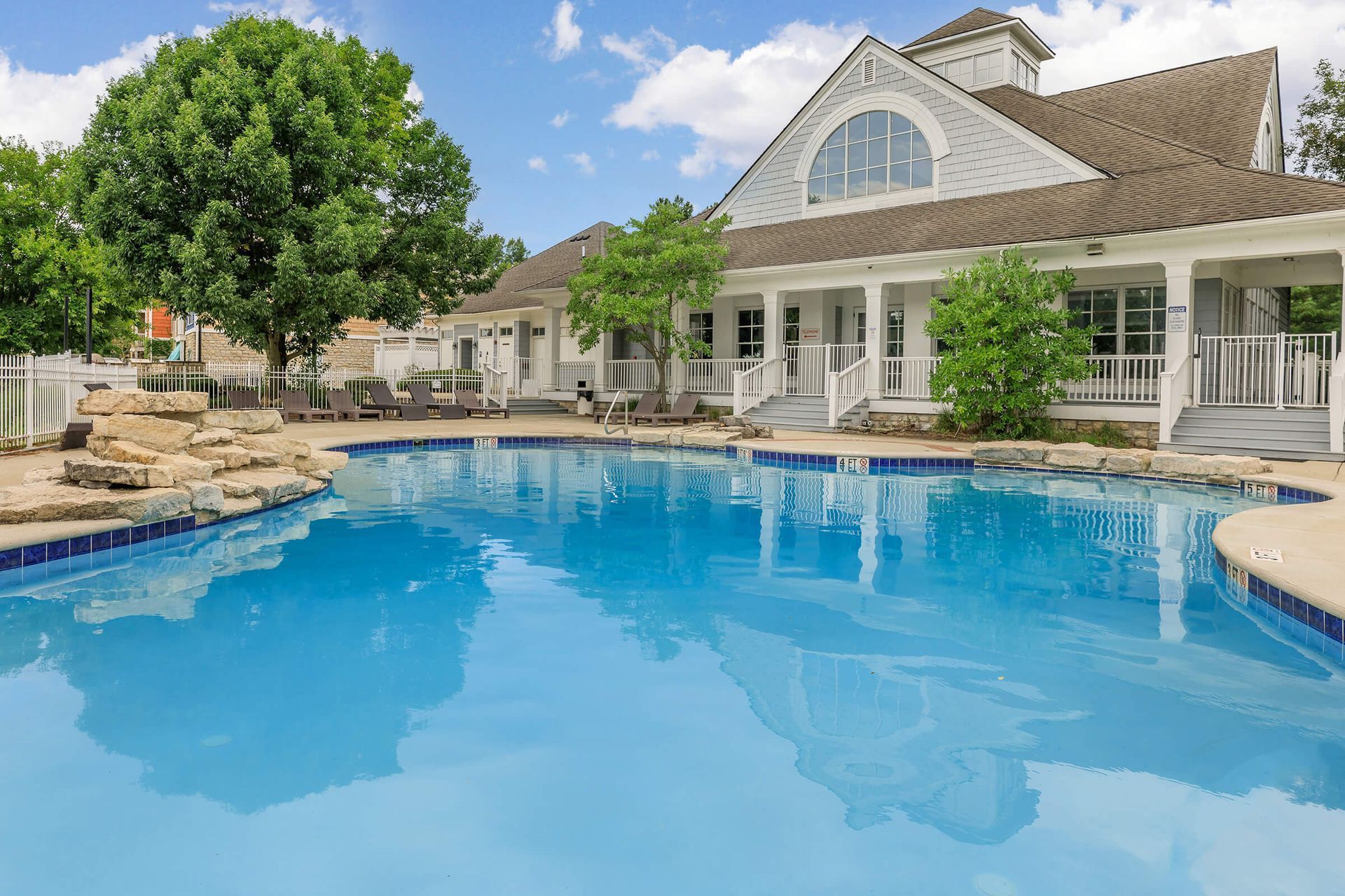 Luxury backyard pool beside a large white house with stone patio, lounge chairs, and blue sky.
