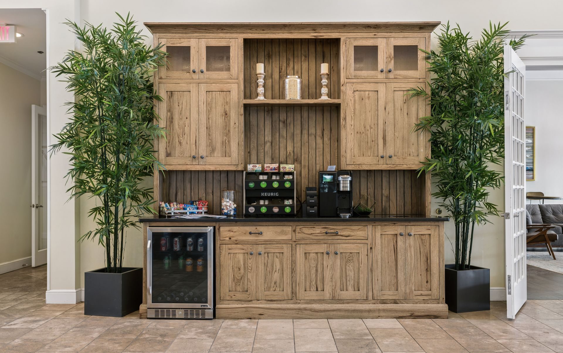 Wooden beverage station with coffee machines, mini fridge, and two tall potted plants in a lobby.