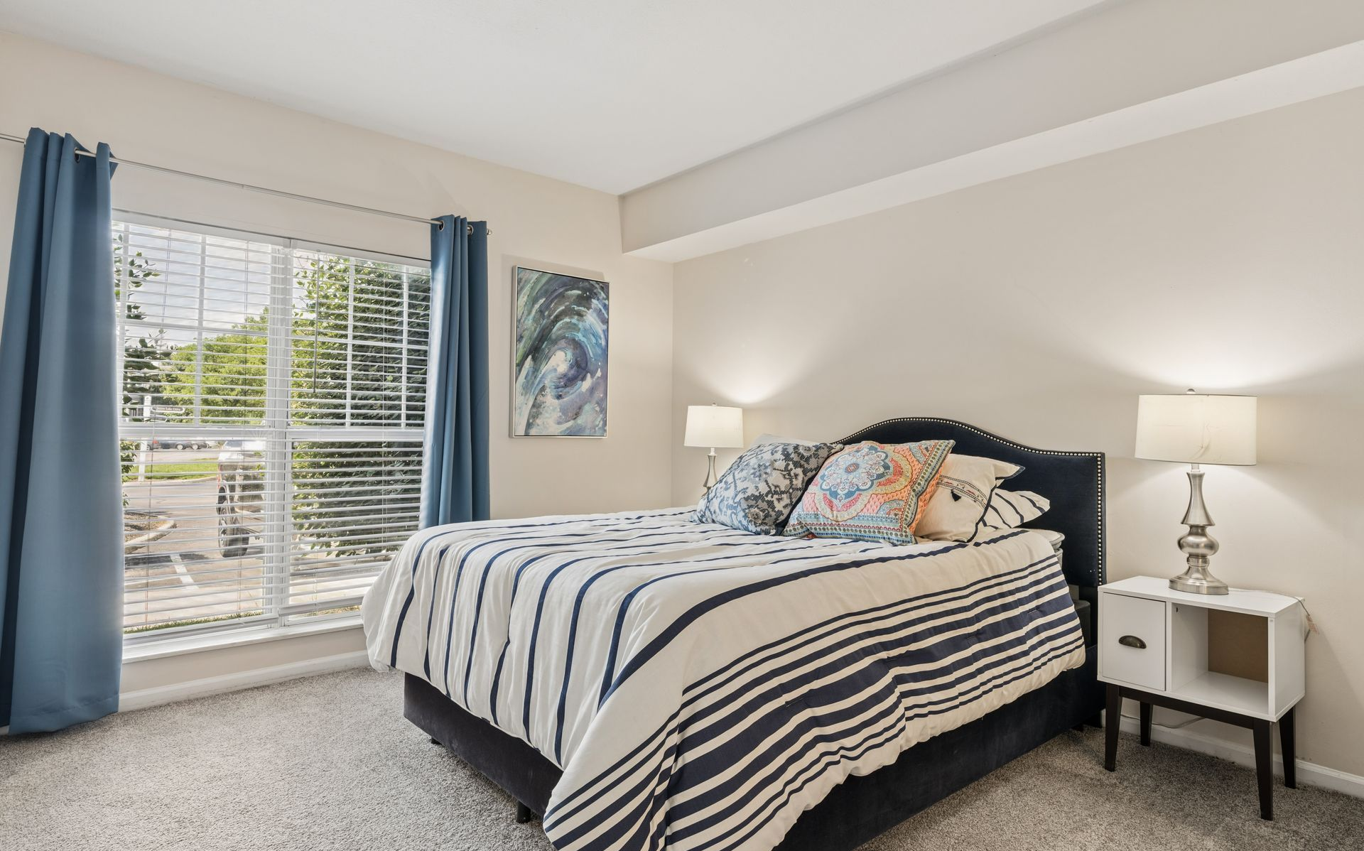 Bright bedroom with striped bedspread, bedside lamps, and large window with blue curtains.