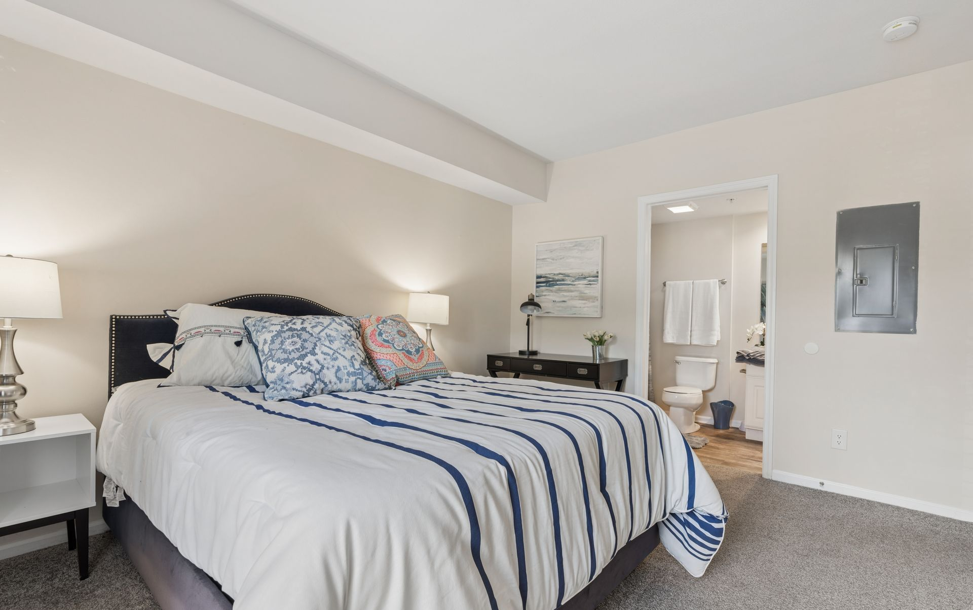 Bedroom with a neatly made bed, striped blanket, bedside lamps, and open doorway to a bathroom.