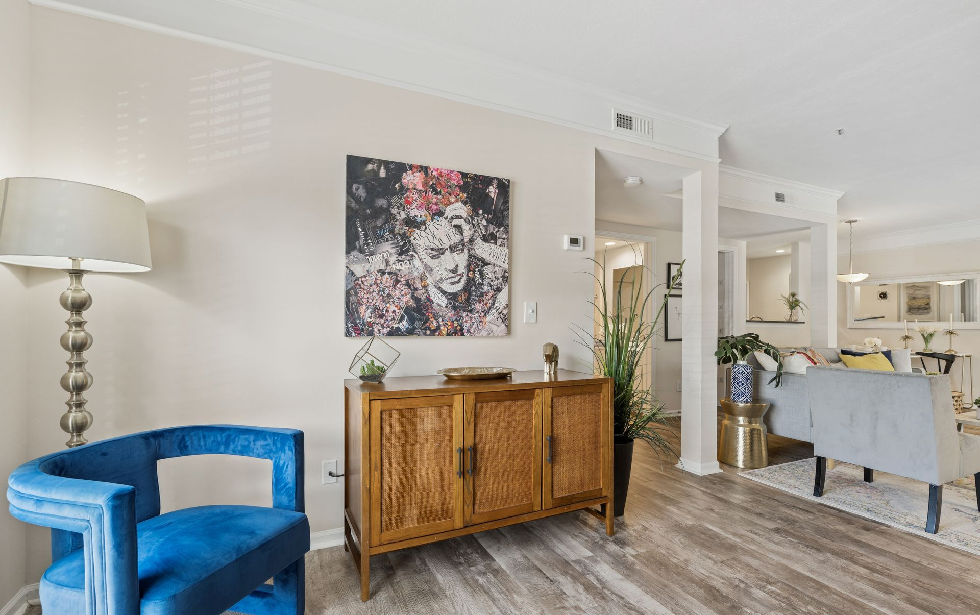 Bright living room with blue chair, wooden sideboard, floral art, and open view to a kitchen and dining area.