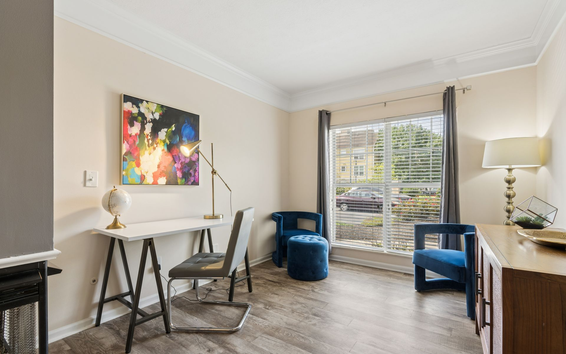 Modern living room with desk, blue chairs, colorful wall art, and large window overlooking greenery.