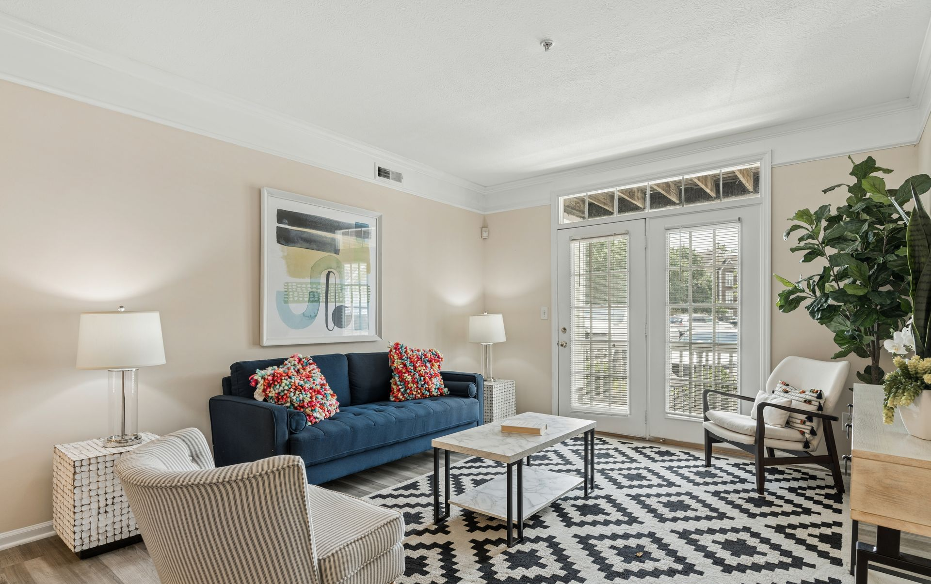 Bright living room with blue sofa, patterned rug, white chairs, and French doors to greenery.