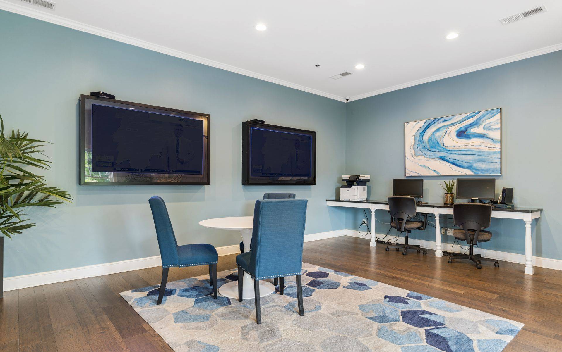 Modern business office with desks, dual monitors, blue chairs, wall-mounted TVs, and a blue geometric rug.
