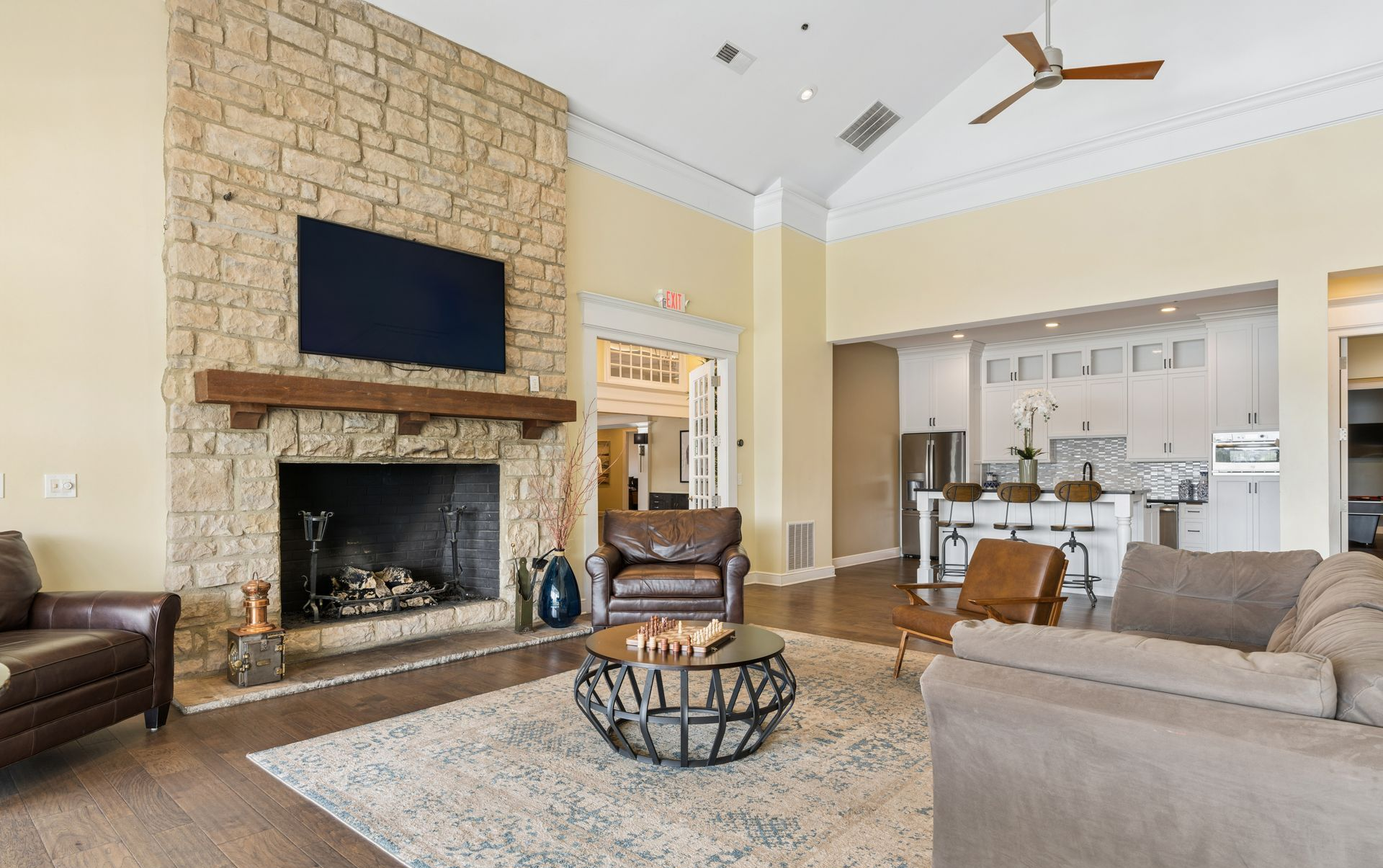 Bright club room with stone fireplace, TV, beige sofas, and open kitchen in the background.