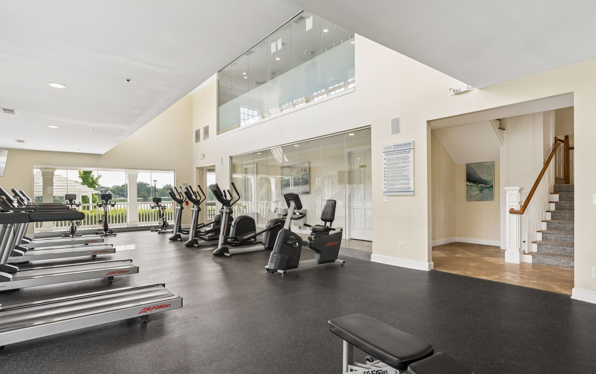 Bright gym with treadmills, ellipticals, weights, and a stairway in the background.