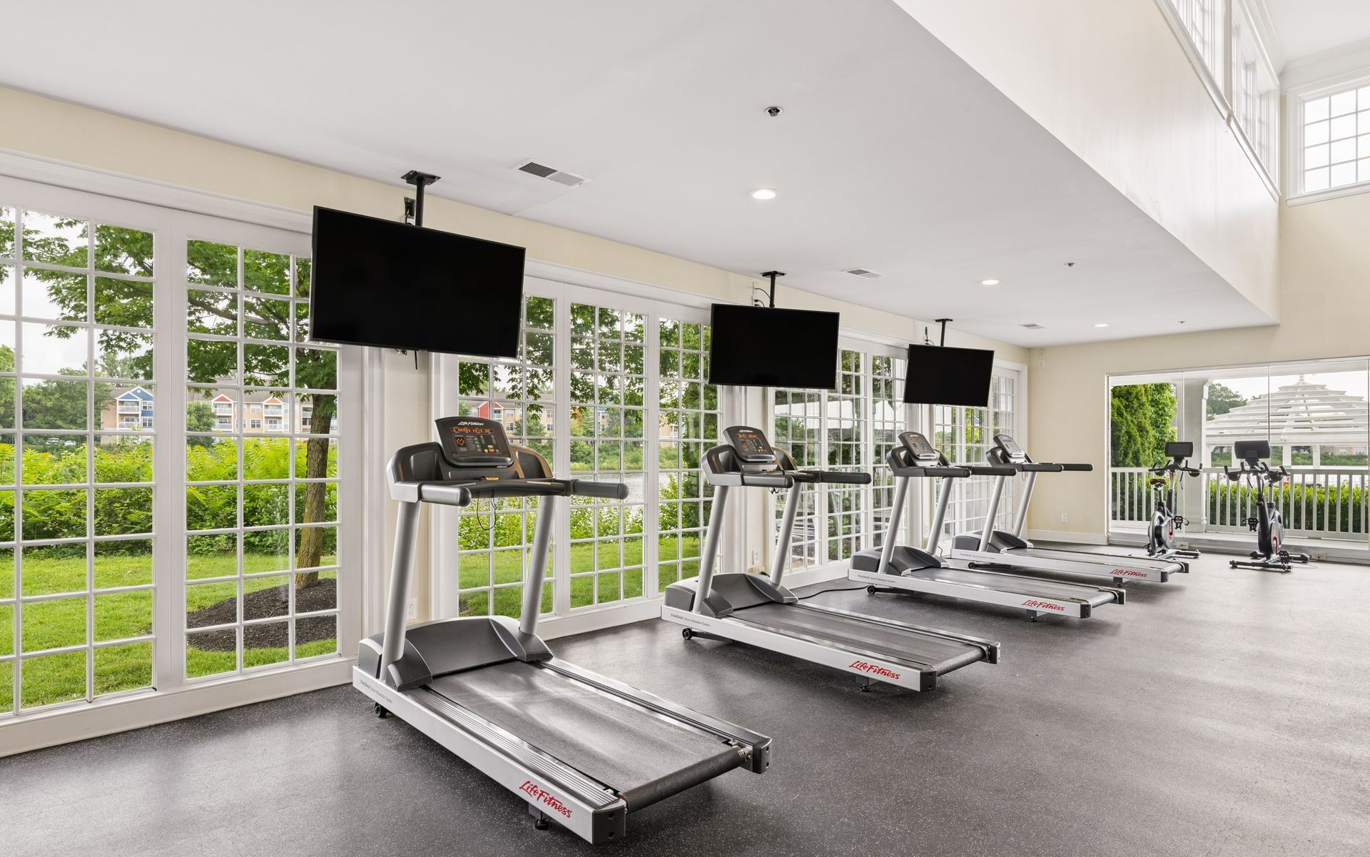 Bright gym with rows of treadmills by large windows overlooking greenery.