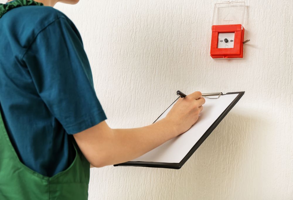 Person in Teal and Green Overalls Inspecting a Red Fire Alarm — Industry Fire Services in Bowen, QLD