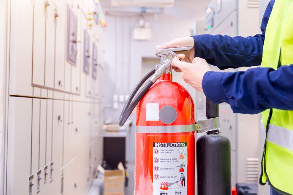 Person in a Yellow Vest Checks a Red Fire Extinguisher Next to Electrical Panels — Industry Fire Services in Bowen, QLD