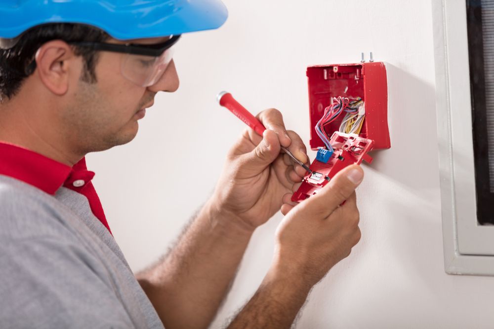 Electrician in Safety Gear Repairs a Red Fire Alarm Panel on a White Wall — Industry Fire Services in Charters Tower, QLD