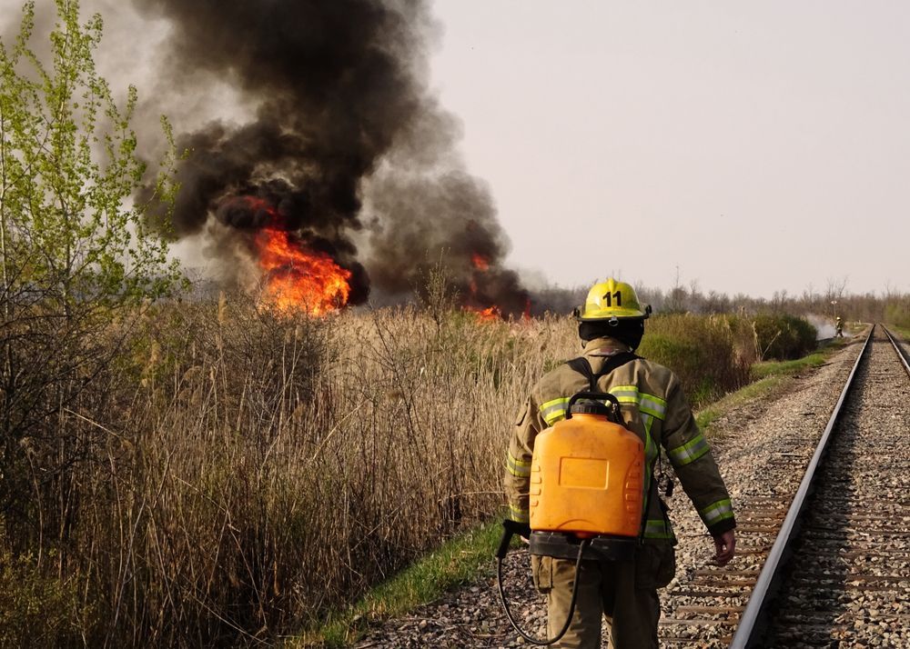 Firefighter With Backpack Sprayer Walks Near Burning Field and Railroad Tracks — Industry Fire Services in Ayr, QLD
